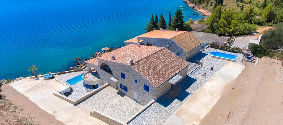 Blick von oben auf das Grundstück und das Haus der Villa Bavaria mit Parkmöglichkeiten direkt am Haus. Dahinter der weite Blick auf die grüne Küste und das blaue Meer.