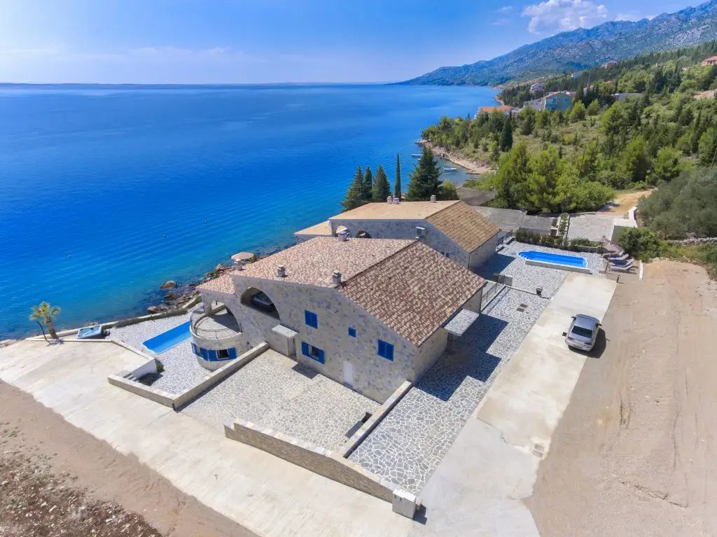 Blick von oben auf das Grundstück und das Haus der Villa Bavaria mit Parkmöglichkeiten direkt am Haus. Dahinter der weite Blick auf die grüne Küste und das blaue Meer.